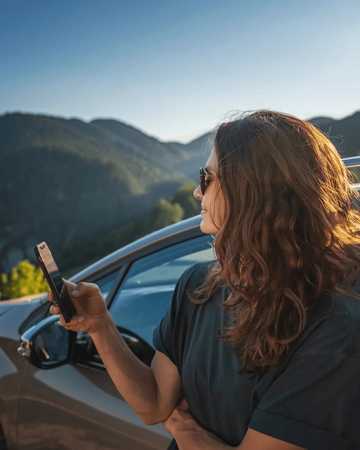 imagen de una chica viendo el movil y que tiene como fondo unas montañas y un coche que ha alquilado a través del servicio de alquiler de coche en asturias alvemaco rent